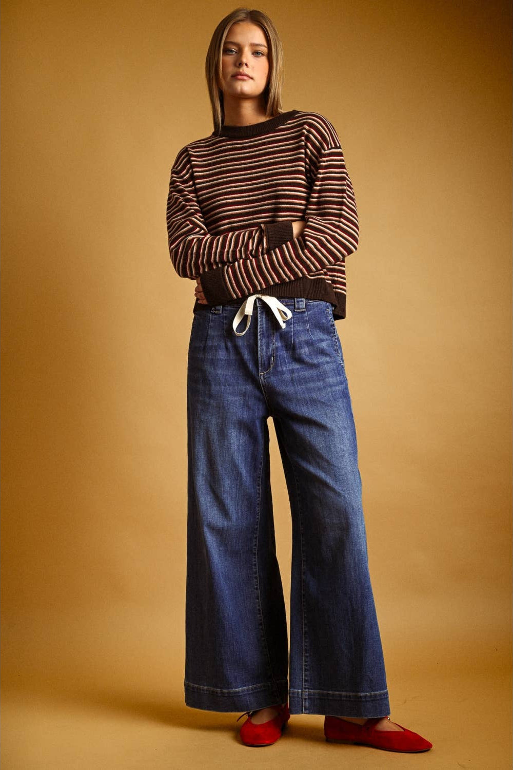 A model stands against a warm tan studio backdrop wearing a brown and multi striped long-sleeve knit top, wide-leg blue jeans with a white drawstring, and red flats.
