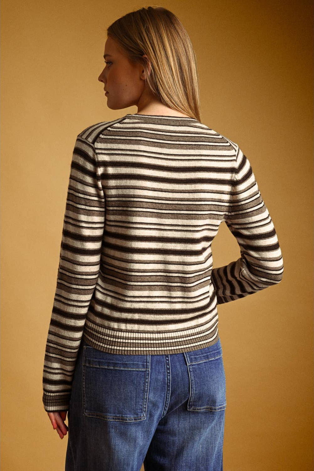 A woman shown from the back wearing a long-sleeve knit sweater with horizontal brown and cream stripes, paired with blue jeans, against a solid warm brown background.