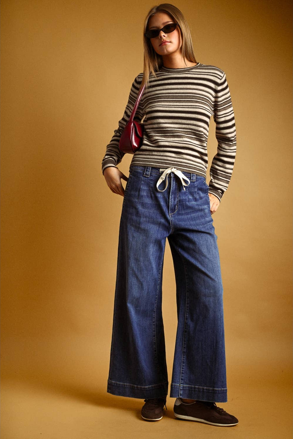 A woman modeling a brown multi striped long-sleeve knit top and wide-leg blue jeans against a warm tan studio backdrop. She is wearing dark sunglasses, a small red shoulder bag, and dark brown shoes.