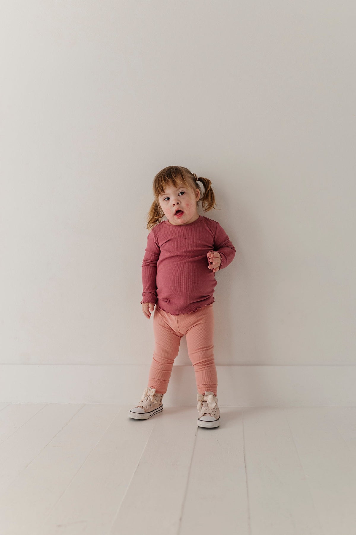 A toddler girl stands against a plain off-white wall wearing a dusty rose long-sleeve top, peach-pink leggings, and light-colored sneakers.