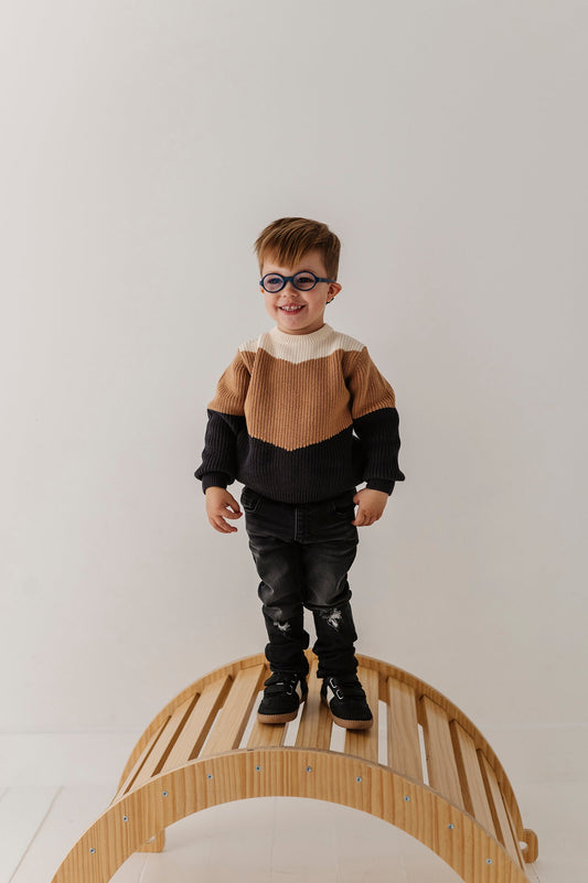 Child standing on a wooden climbing toy in a black, camel and beige sweater and black jeans against a white wall