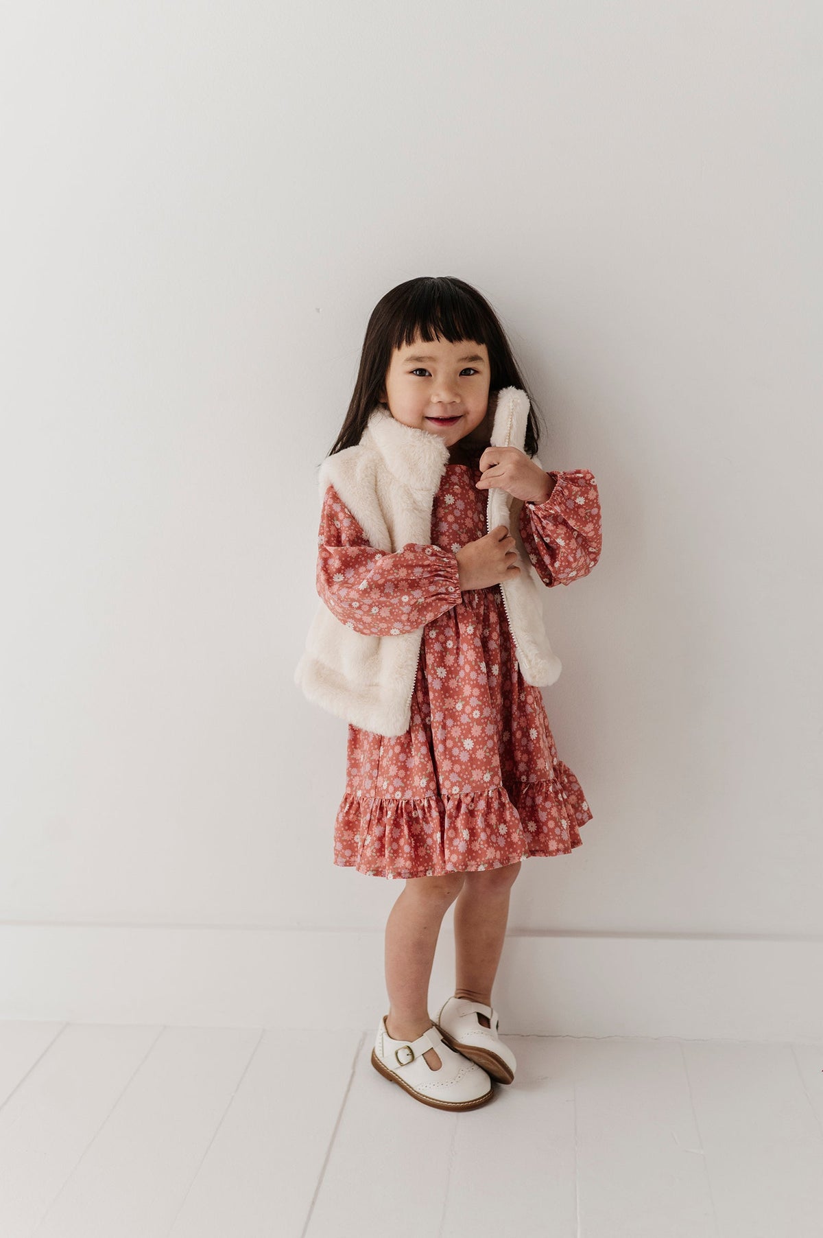 A young girl with dark hair stands against a white wall wearing a pink and white floral long-sleeve dress with a fluffy cream faux-fur vest and white Mary Jane shoes.