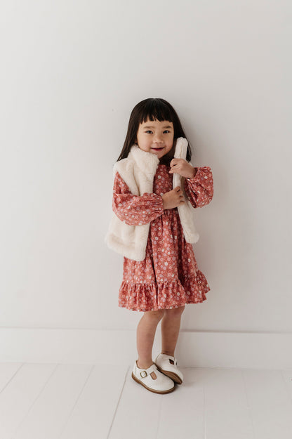 A young girl with dark hair stands against a white wall wearing a pink and white floral long-sleeve dress with a fluffy cream faux-fur vest and white Mary Jane shoes.