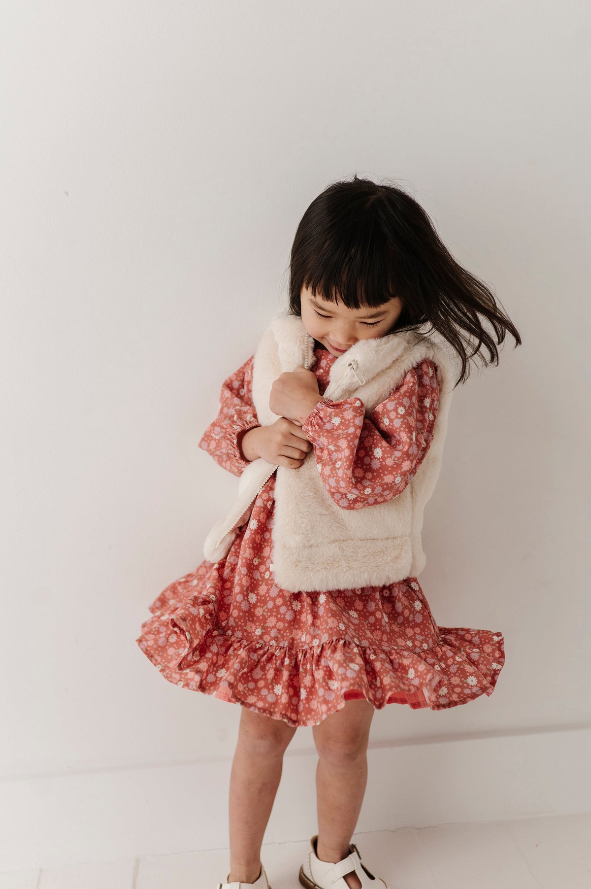 Child wearing a floral dress and cream fur vest against a plain background