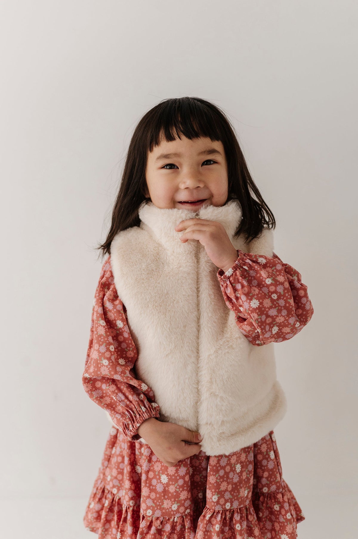 Young girl wearing a cream fur vest over a red floral dress on a plain background