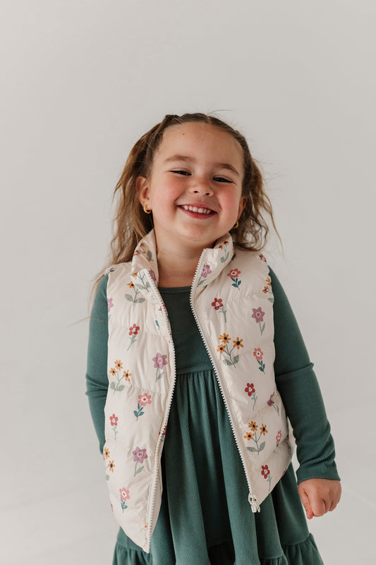 Young girl wearing a floral embroidered puffer vest over a green dress on a plain background