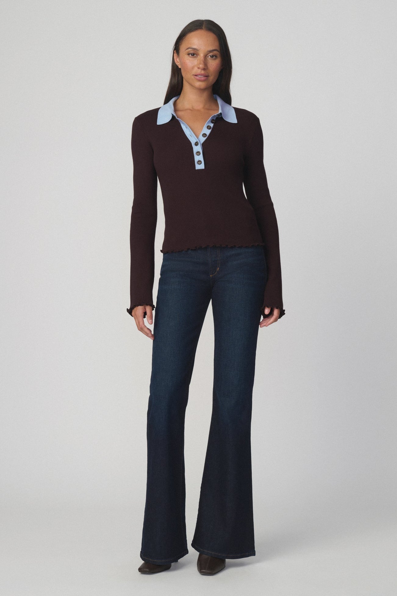Woman wearing an espresso colored polo style sweater with a light blue collar and dark flare blue jeans on a light gray background.