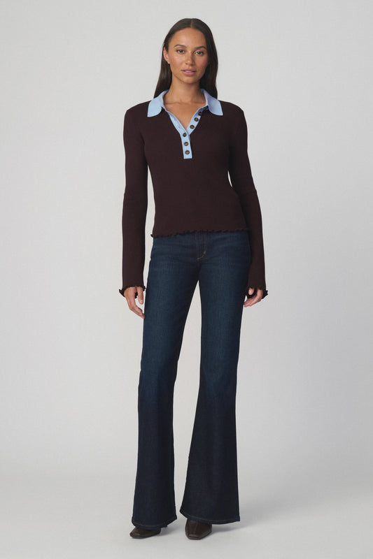 Woman wearing an espresso colored polo style sweater with a light blue collar and dark flare blue jeans on a light gray background.