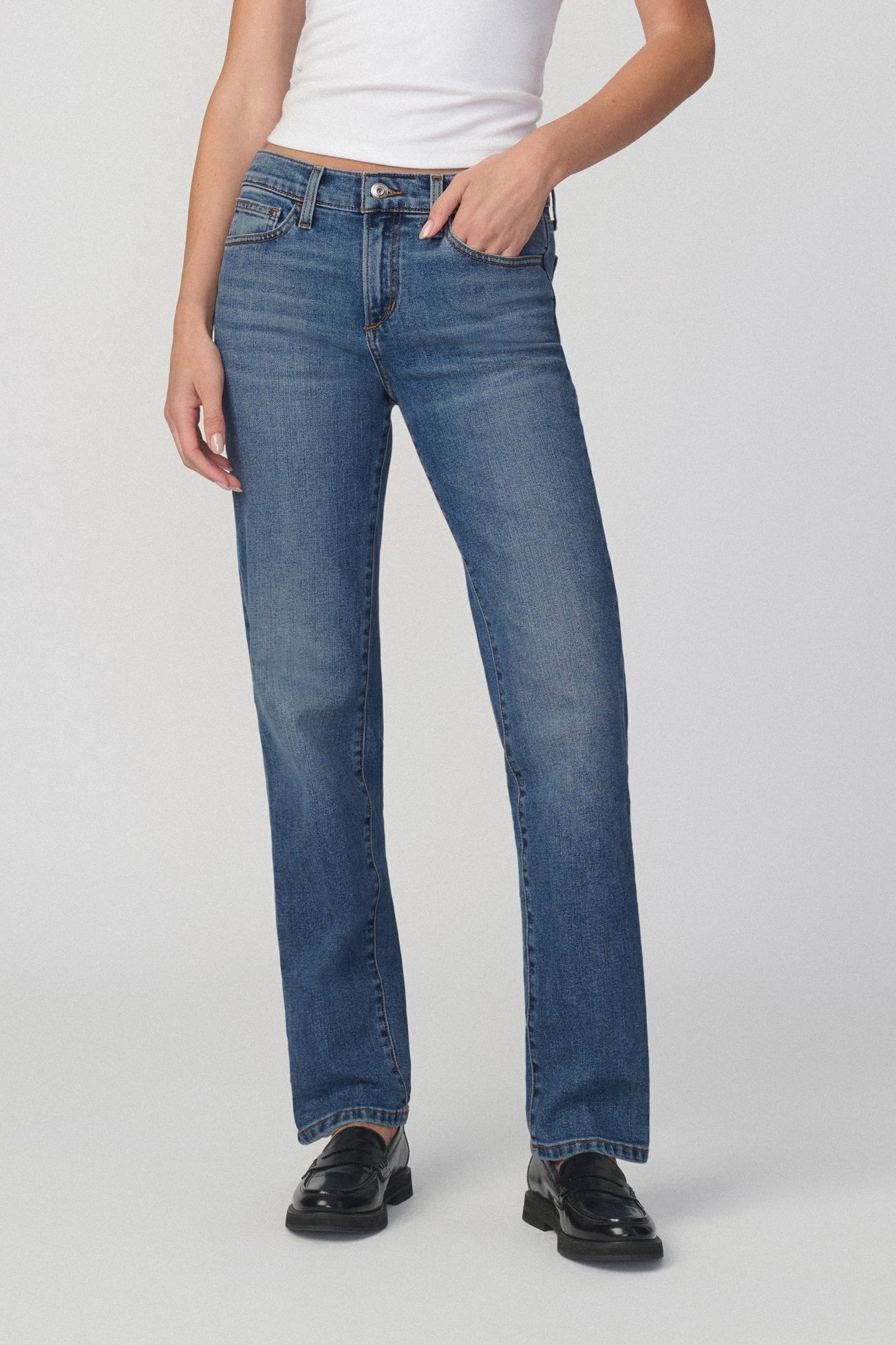 A model wearing a pair of medium-wash amalfi blue straight-leg jeans, styled with a white top and black leather loafers, photographed against a neutral light gray studio background.
