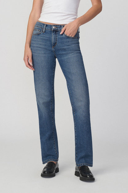A model wearing a pair of medium-wash amalfi blue straight-leg jeans, styled with a white top and black leather loafers, photographed against a neutral light gray studio background.