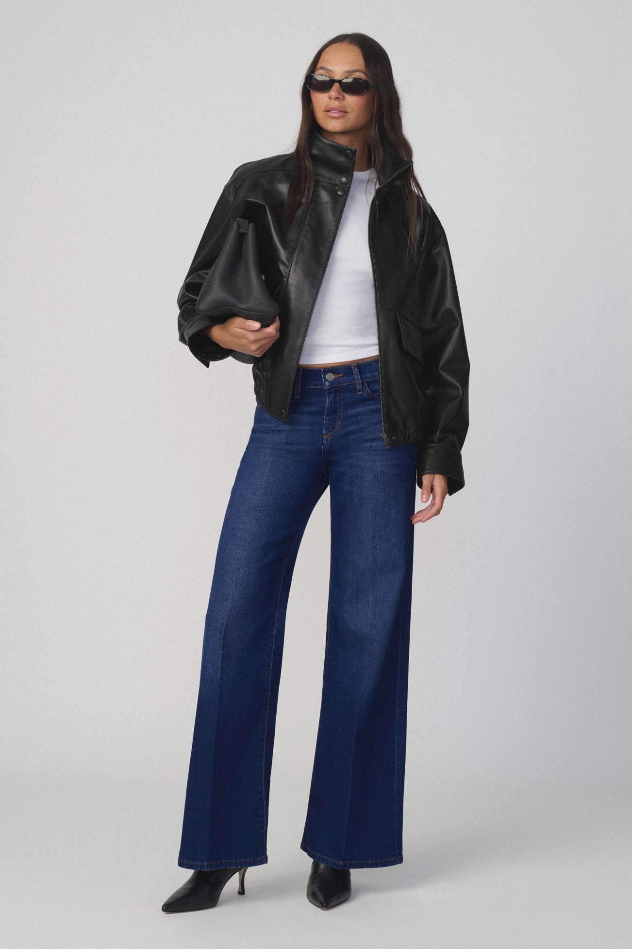 Woman wearing a black leather jacket, white shirt, and The lou lou dark blue jeans in splendor color on a gray background