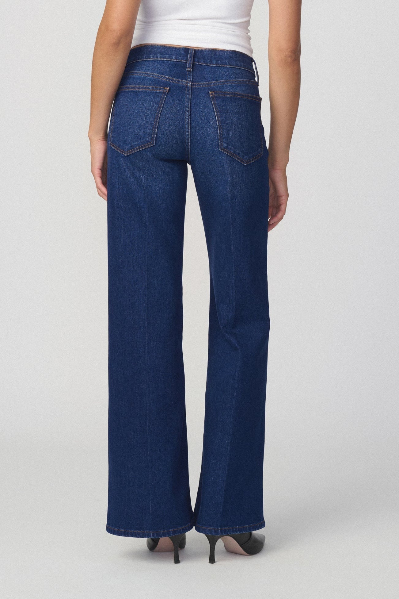 Back view of woman wearing the lou lou dark blue jeans in splendor color with heels on a plain background