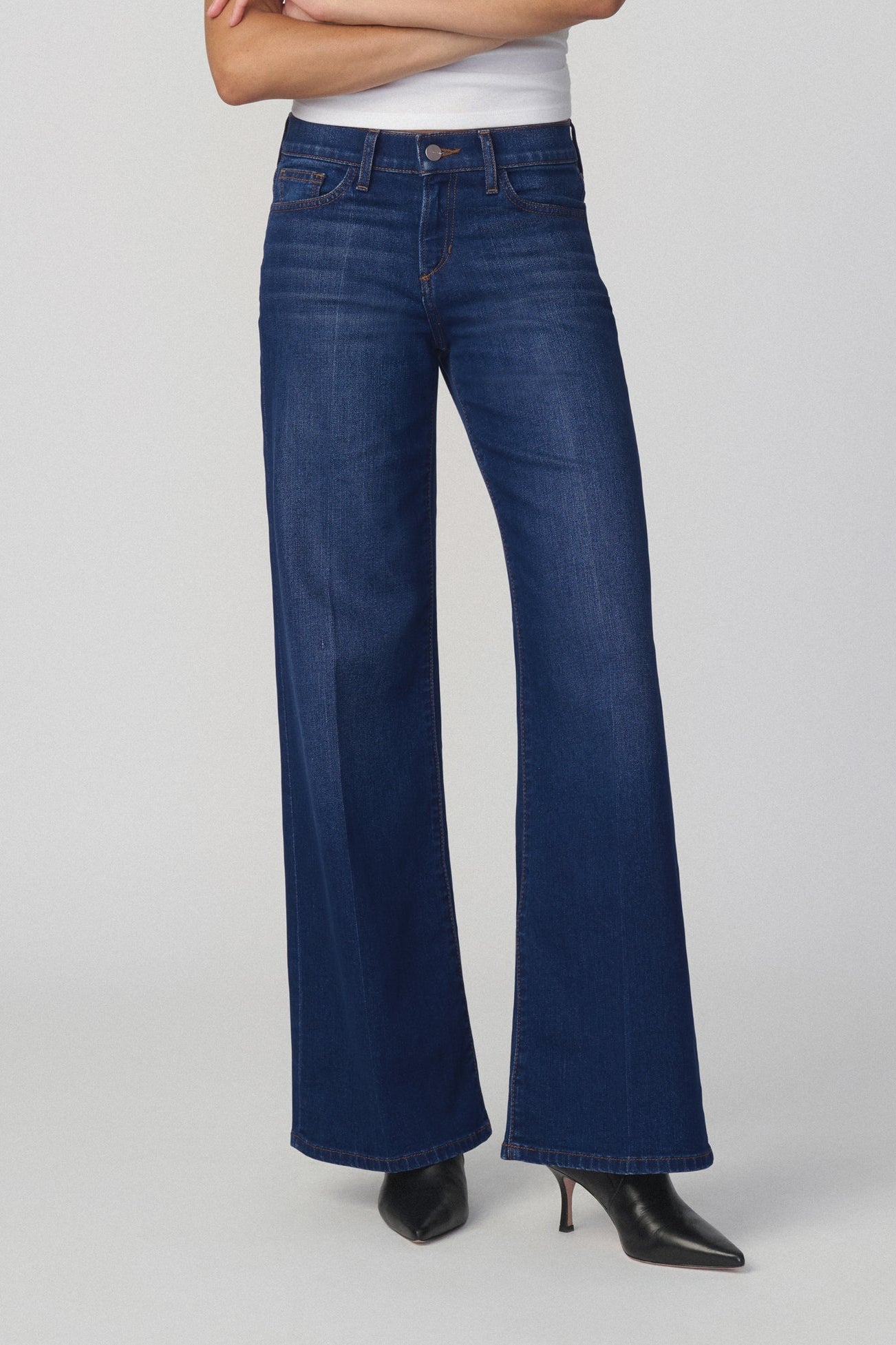 A model wearing a pair of dark blue wide-leg lou lou flare jeans in color splendor, styled with a white top and black pointed-toe heels, photographed against a neutral light gray studio background.