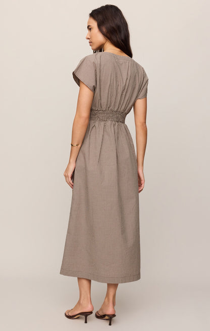 Back view of woman wearing a dark chocolate colored Sanna gingham dress against a plain background