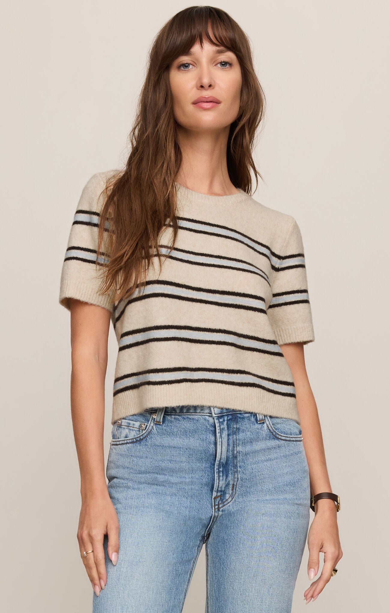 Front view of woman wearing a light oatmeal heather  striped Gatewood sweater and blue jeans on a plain background