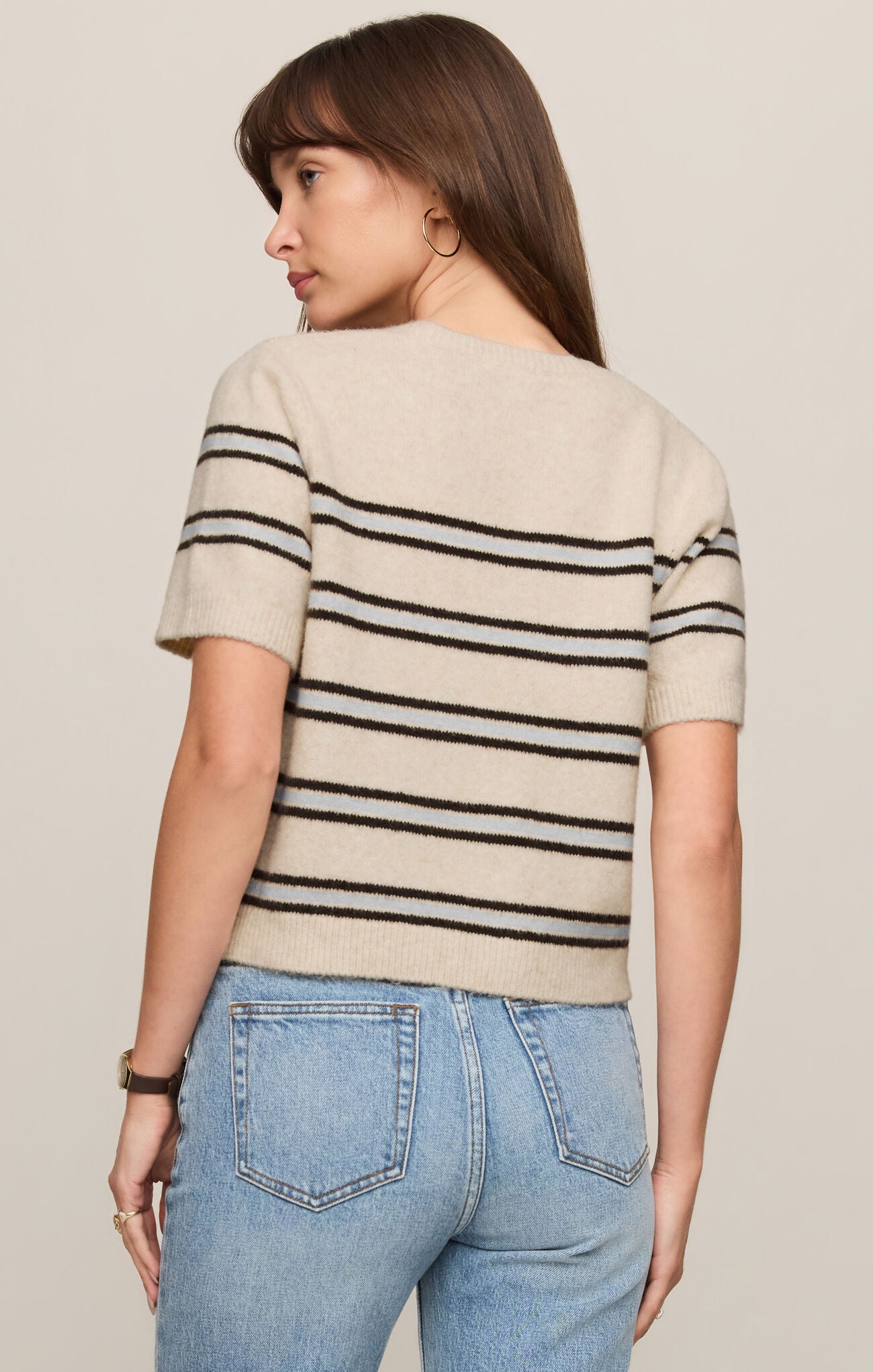 Woman wearing a light oatmeal heather  striped Gatewood sweater and blue jeans on a plain background