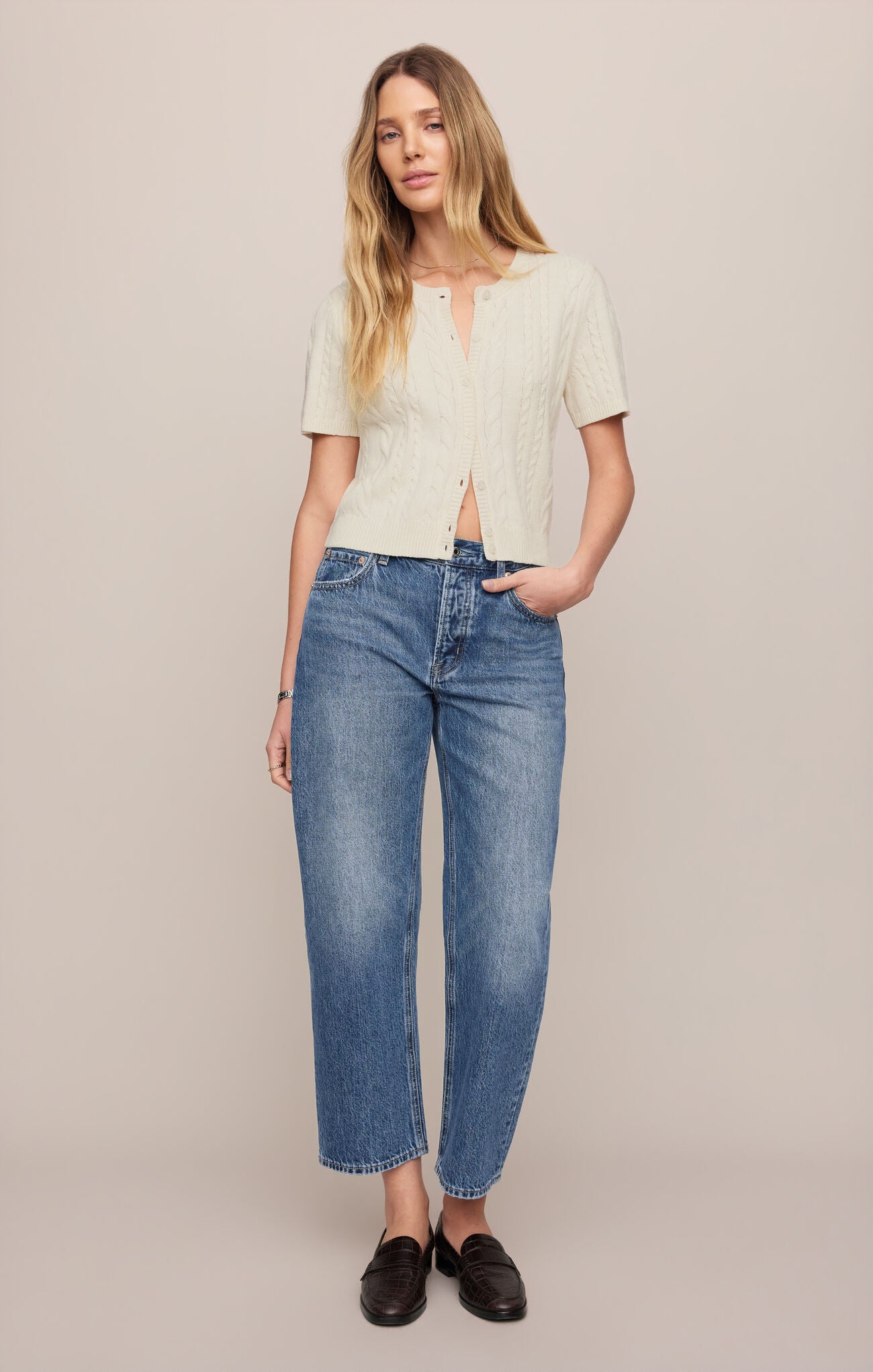 Woman wearing a beige top and breakpoint colored off-duty bowed ankle jeans against a plain background