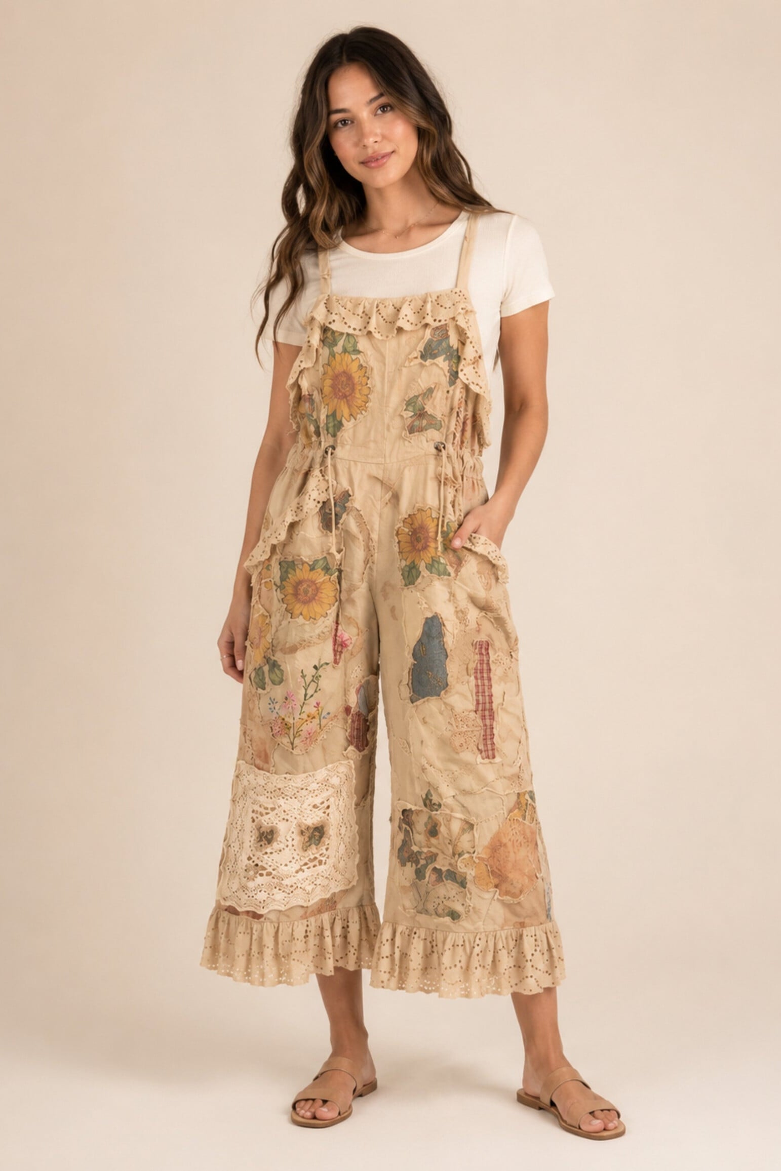 A woman modeling a beige patchwork wide-leg overall jumpsuit with floral meadows print, lace trim, and ruffled details, styled over a white t-shirt and sandals against a solid beige background.