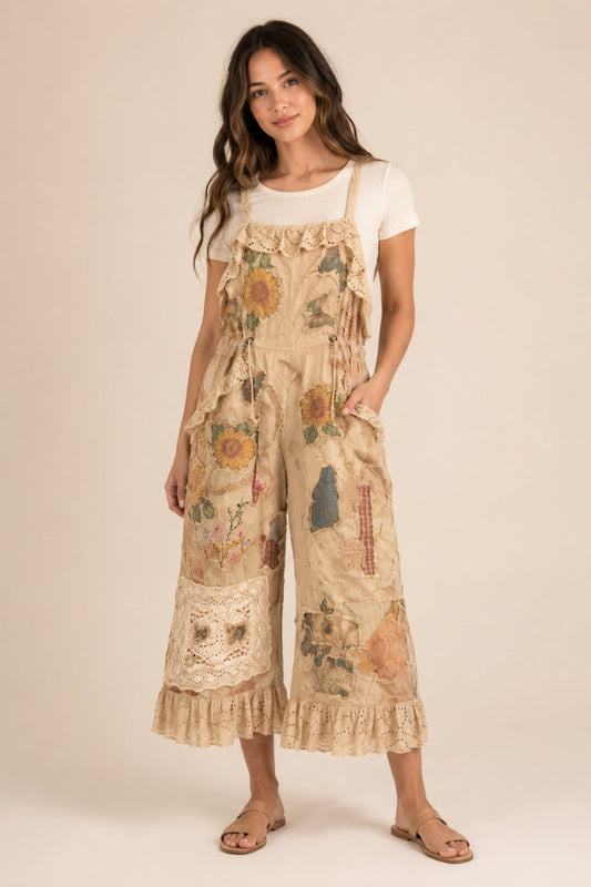 A woman modeling a beige patchwork wide-leg overall jumpsuit with floral meadows print, lace trim, and ruffled details, styled over a white t-shirt and sandals against a solid beige background.