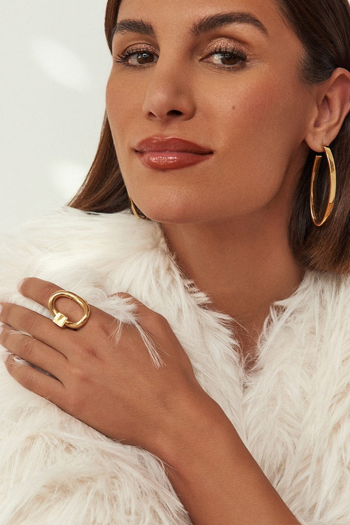 A close-up lifestyle shot of a woman wearing gold jewelry, featuring a large gold hoop earring and a chunky gold Queen ring on her finger, styled with a white faux fur coat against a clean white background.