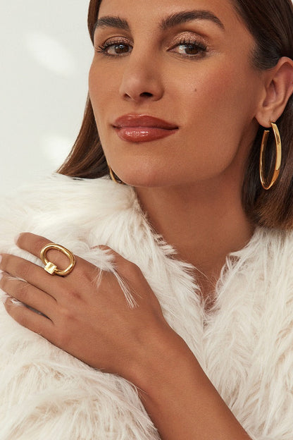 A close-up lifestyle shot of a woman wearing gold jewelry, featuring a large gold hoop earring and a chunky gold Queen ring on her finger, styled with a white faux fur coat against a clean white background.