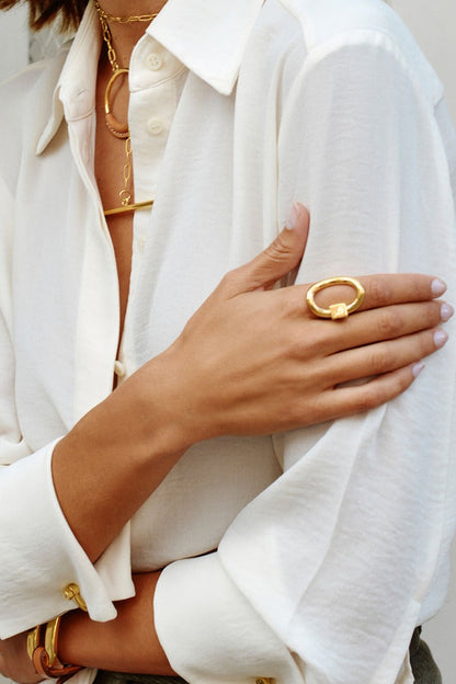 A close-up of a person wearing a white button-up shirt and layered gold jewelry, including a chunky gold Queen ring on one hand.
