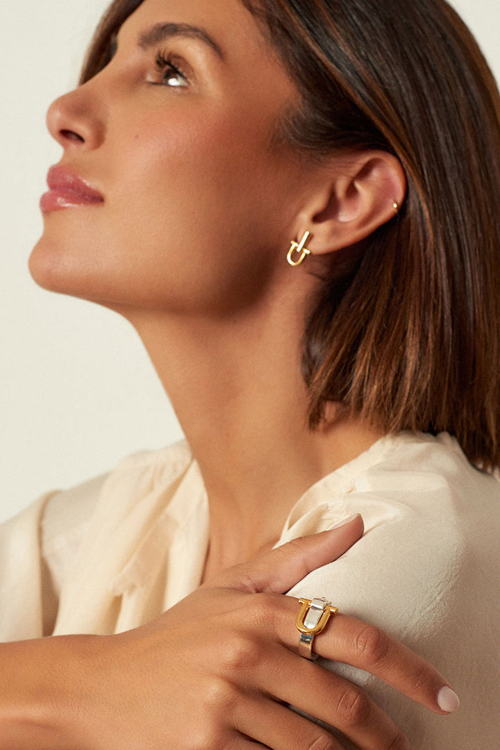 A close-up lifestyle shot of a woman wearing a gold and silver Shackle ring with a rectangular shape and a small gold hoop earring, looking upward against a warm off-white background.