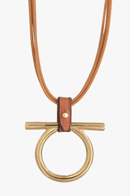 A modern Omega necklace featuring a gold-tone ring and bar pendant with a small camel brown leather accent, suspended from a multi-strand camel colored cord, shown against a light gray background.