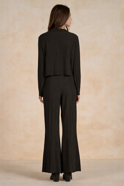 A woman viewed from the back wearing a black pleated long-sleeve cropped jacket and matching pleated wide-leg trousers against a warm beige textured studio backdrop.
