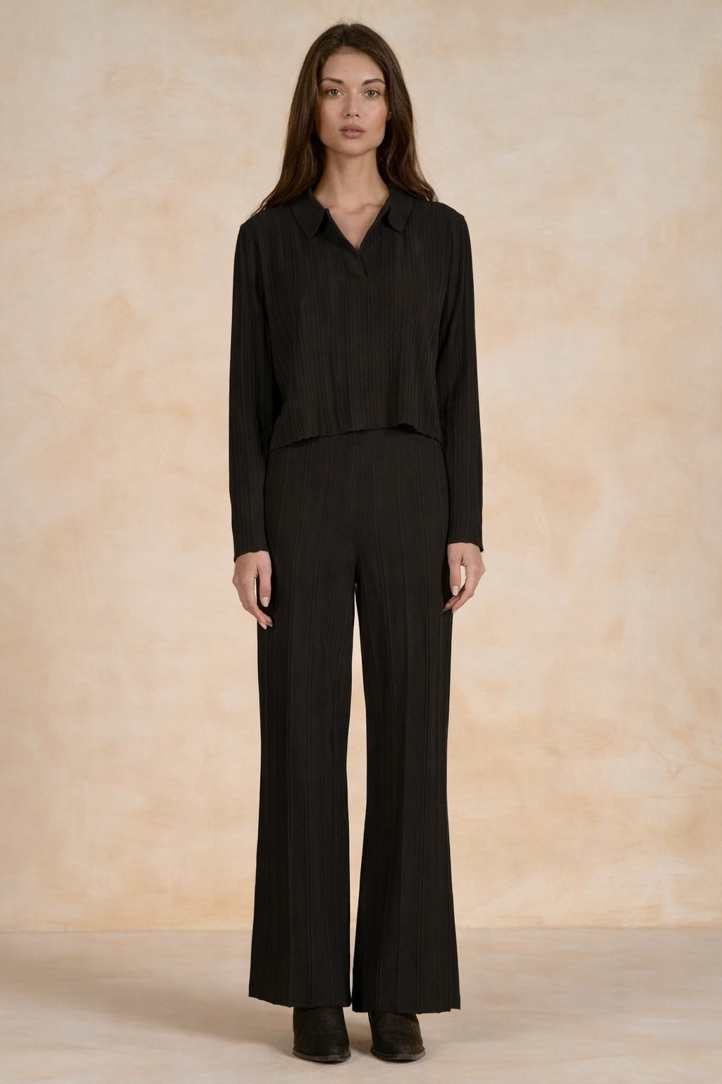 A woman stands facing forward wearing a black long-sleeve collared top and matching black wide-leg pants against a warm beige textured studio backdrop.