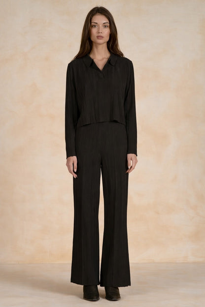 A woman stands facing forward wearing a black long-sleeve collared top and matching black wide-leg pants against a warm beige textured studio backdrop.