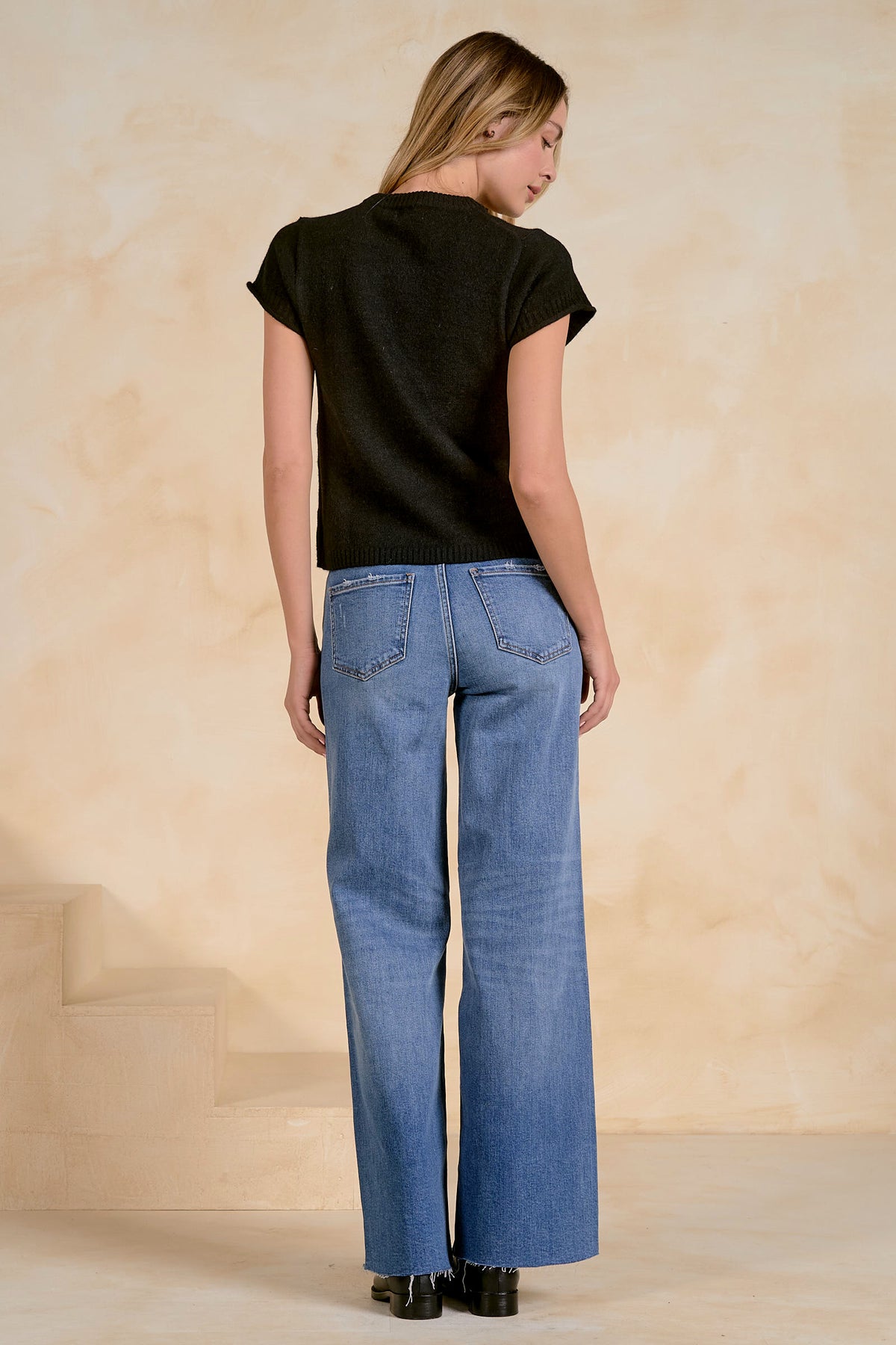A woman is shown from behind wearing a black short-sleeve sweater and wide-leg blue jeans, standing against a textured beige wall.