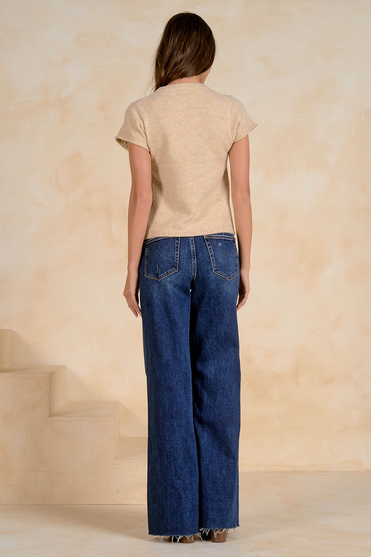 A woman shown from the back wearing a stone colored short-sleeve sweater and wide-leg blue denim jeans, standing against a textured beige wall.