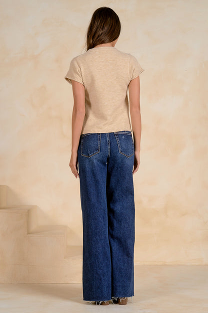 A woman shown from the back wearing a stone colored short-sleeve sweater and wide-leg blue denim jeans, standing against a textured beige wall.