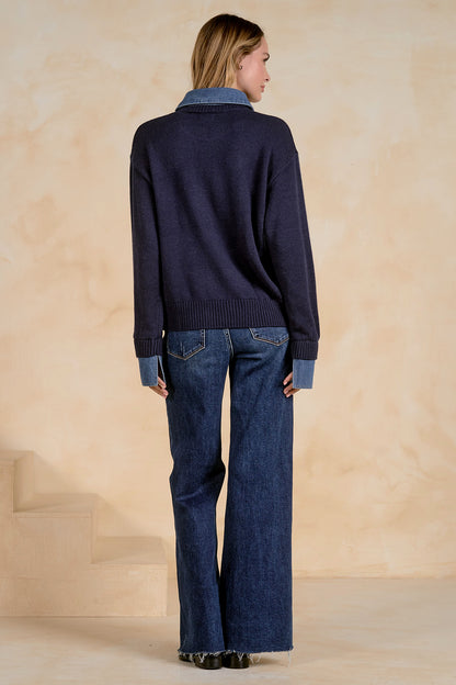 A woman is shown from the back wearing a navy blue knit sweater with light blue ribbed denim collar, cuffs, and hem, paired with dark blue wide-leg jeans, against a warm beige textured studio backdrop.