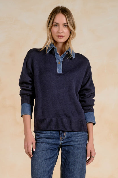 A woman modeling a navy blue knit sweater with a light blue denim collar, paired with blue jeans, against a warm beige textured background.