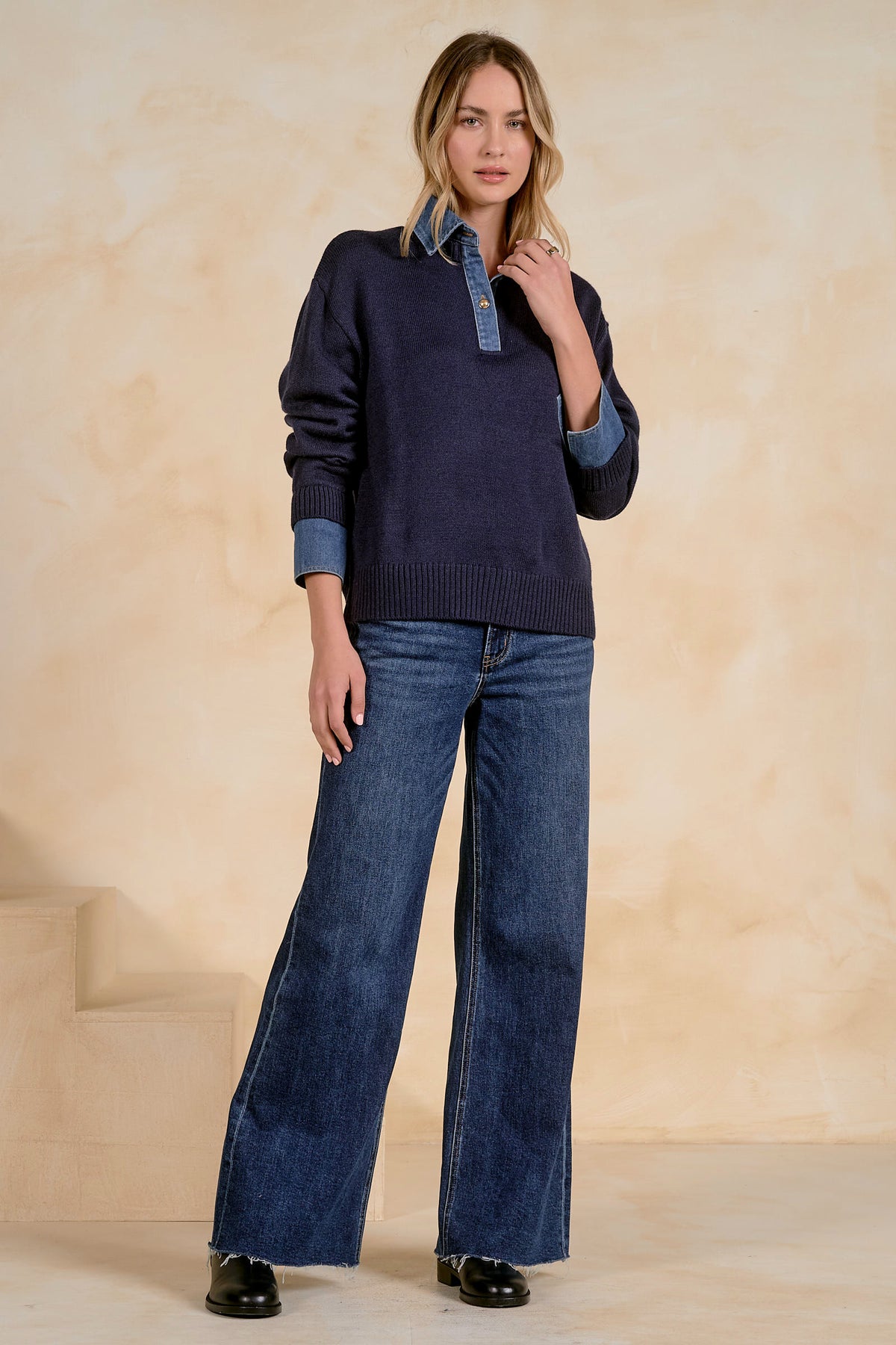 A woman modeling a dark navy blue knit sweater with contrasting light blue denim collar and cuffs, paired with wide-leg blue jeans and black shoes, set against a warm beige textured studio backdrop.