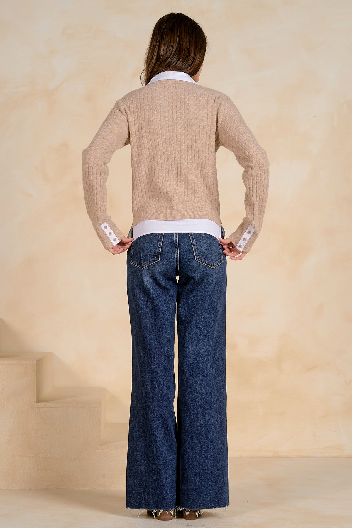 Person wearing a tan cardigan with a white collar and bottom and blue jeans against a beige background
