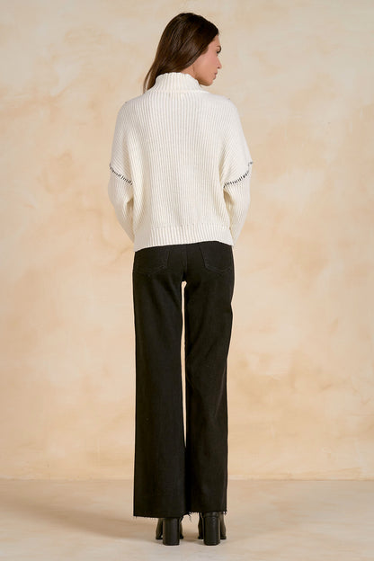 Woman wearing an off white sweater and black pants against a beige background