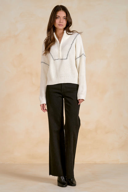 Woman wearing an off white quarter zip sweater with black accents and black pants against a beige background
