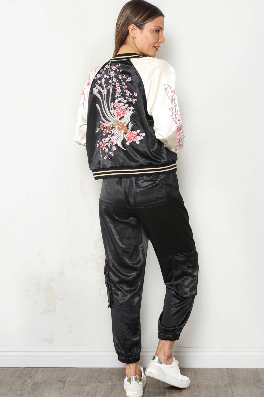 A woman stands with her back to the camera wearing a black satin bomber jacket with white sleeves and pink cherry blossom embroidery, paired with black satin cargo pants and white sneakers, against a white wall.