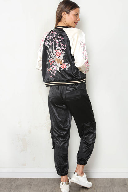 A woman stands with her back to the camera wearing a black satin bomber jacket with white sleeves and pink cherry blossom embroidery, paired with black satin cargo pants and white sneakers, against a white wall.