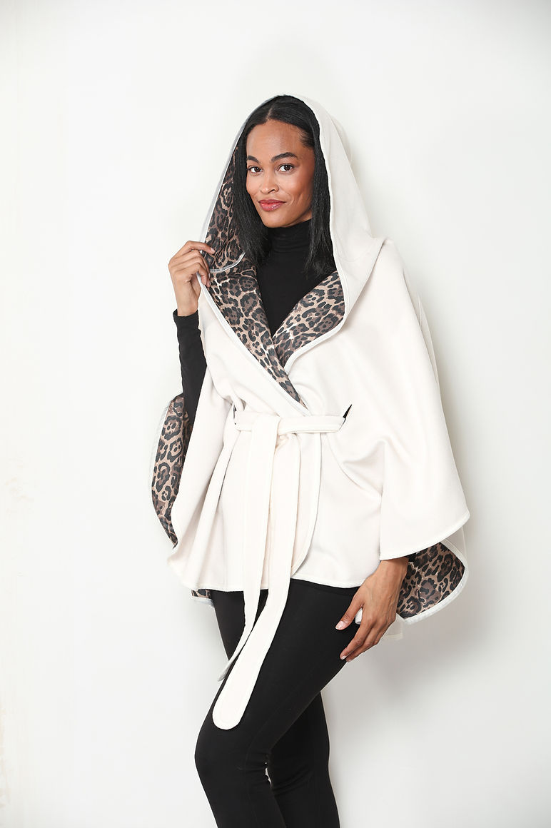 A woman modeling an ivory hooded wrap or poncho-style coat with leopard-print trim and lining, worn over a black turtleneck and black pants, against a plain white studio background.