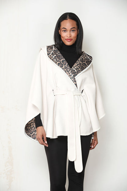 A woman modeling an ivory wrap-style cape jacket with leopard print trim on the collar and sleeve cuffs, worn over a black turtleneck and black pants against a light off-white background.