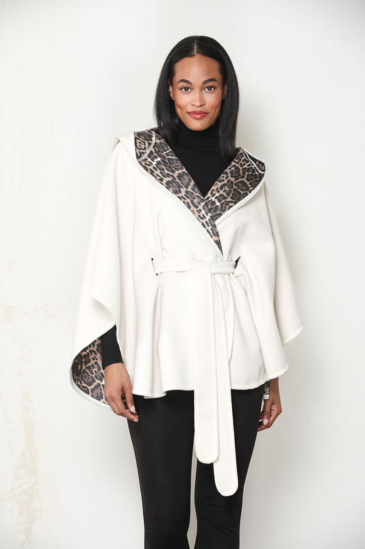 A woman modeling an ivory wrap-style cape jacket with leopard print trim on the collar and sleeve cuffs, worn over a black turtleneck and black pants against a light off-white background.
