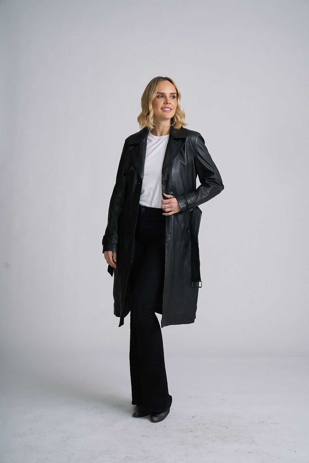 A woman modeling a black leather trench coat over a white top and black pants, standing against a light gray studio background.