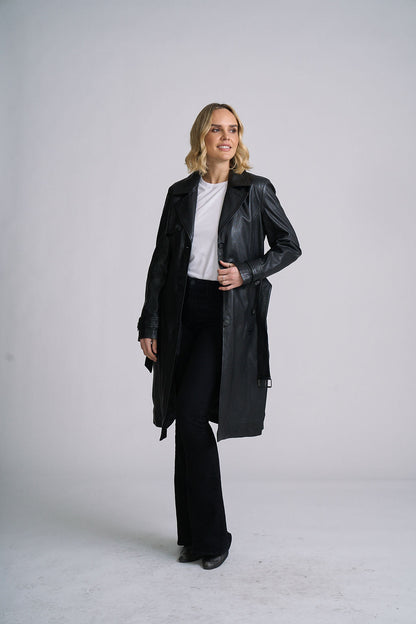 A woman modeling a black leather trench coat over a white top and black pants, standing against a light gray studio background.
