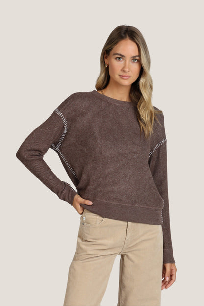 A woman modeling a brown knit sweater with decorative white stitching along the shoulders and sleeves, paired with beige pants, against a solid light gray background.