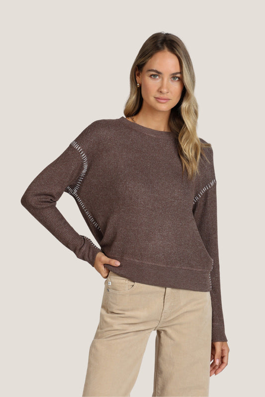 A woman modeling a brown knit sweater with decorative white stitching along the shoulders and sleeves, paired with beige pants, against a solid light gray background.