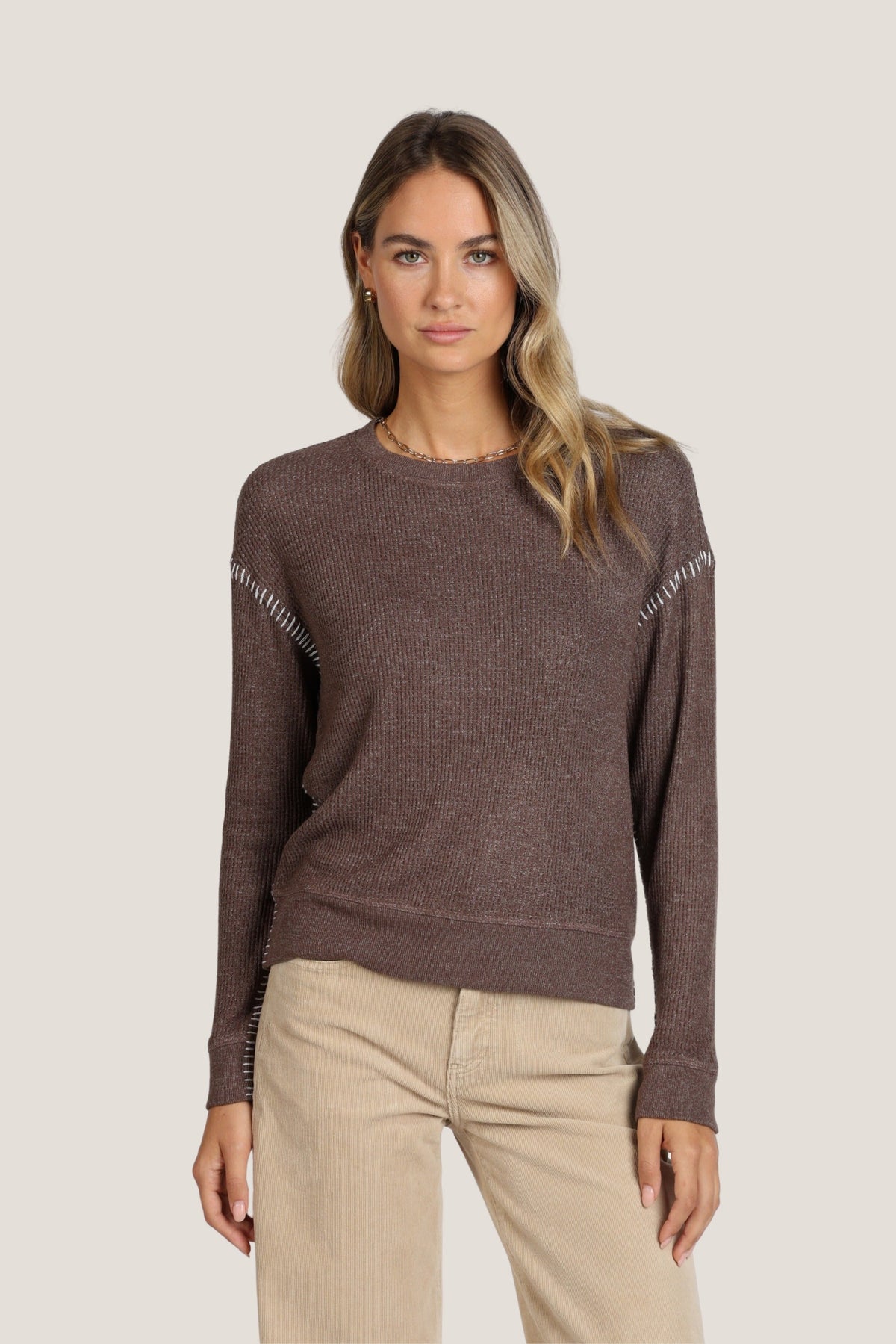 A woman modeling a soft brown long-sleeve knit sweater with textured contrast stitching details on the shoulders and cuffs, paired with beige pants, against a solid light gray background.